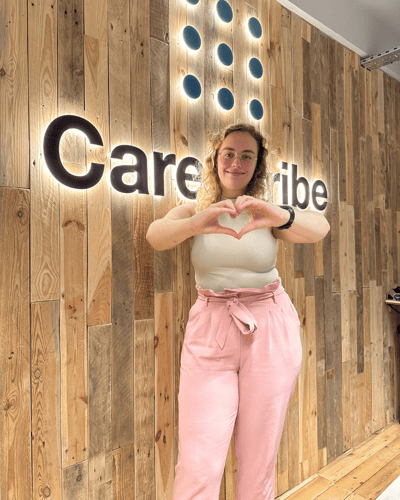 Felicity standing in front of the CareScribe logo in the office with her hands in the shape of a heart, smiling towards the camera